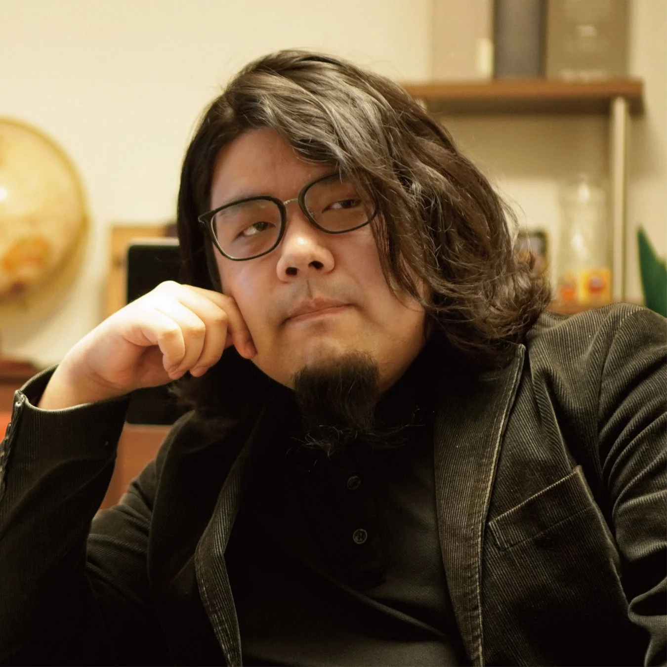 眼鏡と顎髭の男性が、黒いジャケットを着用し、肘をついてカメラを見つめているポートレート写真です。室内で撮影されており、背景には棚や地球儀のようなものが見えます。