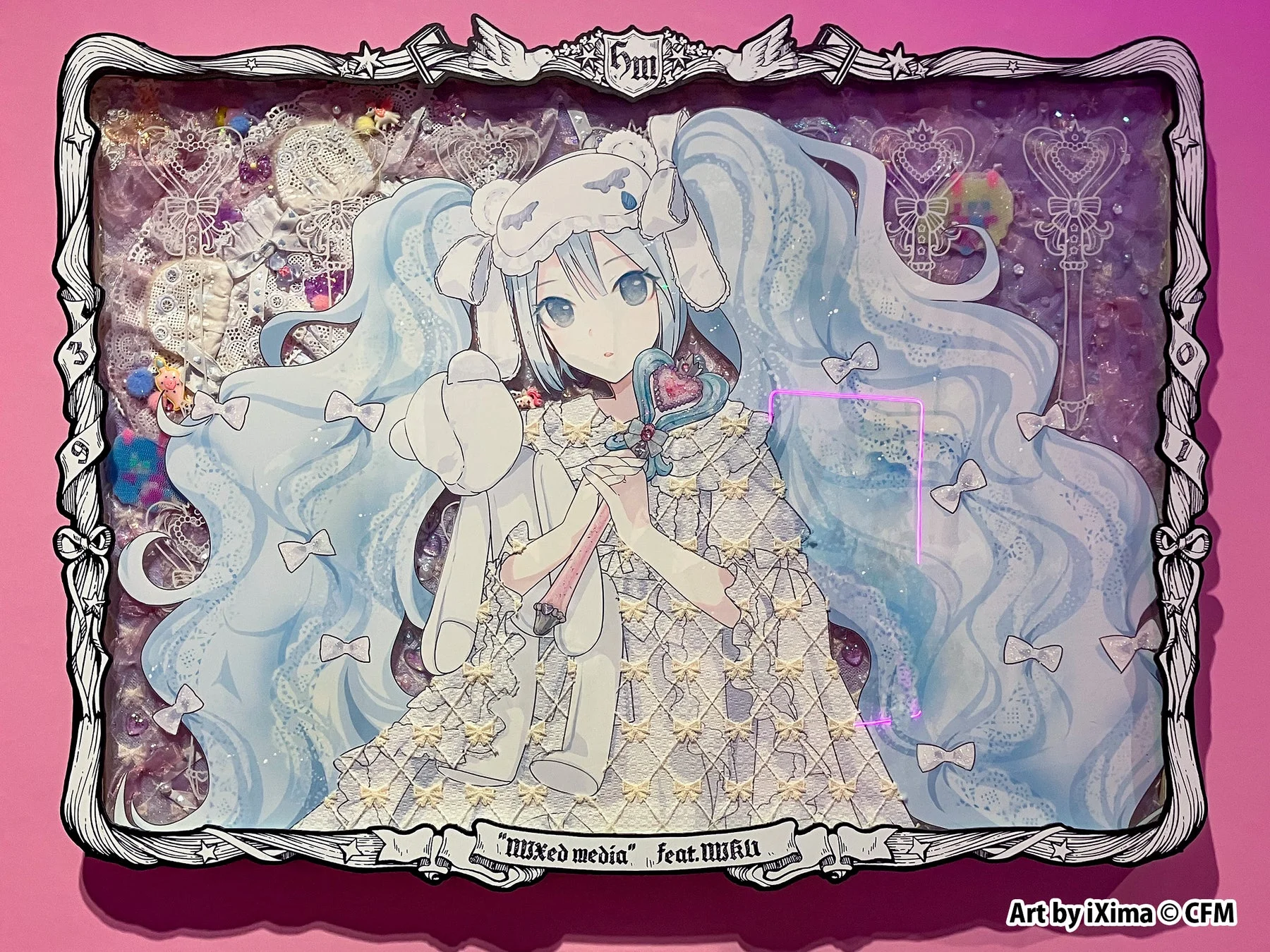 ピンクの壁に飾られた初音ミクのミックスメディアアートです。白いドレスを着たミクがぬいぐるみと魔法の杖を持ち、背景にはレースやリボン、装飾品が詰め込まれたシャドーボックスが特徴的な、夢のような可愛らしい作品です。