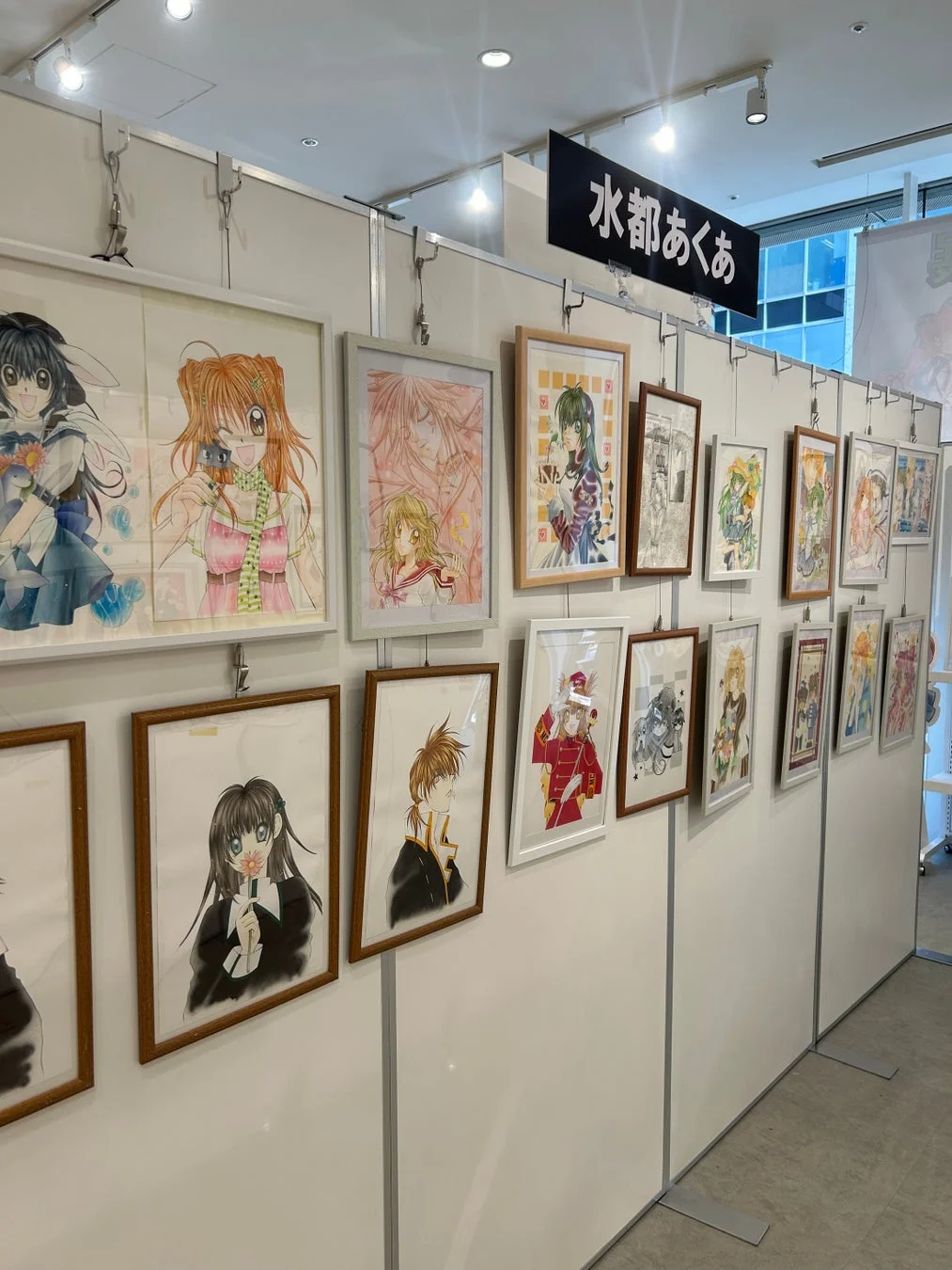 白い壁に多数の額装されたアニメ・漫画風イラストが展示されている様子。主に少女漫画テイストのキャラクターが描かれており、上部には「水都あくあ」と書かれた看板が見える展示会の風景です。