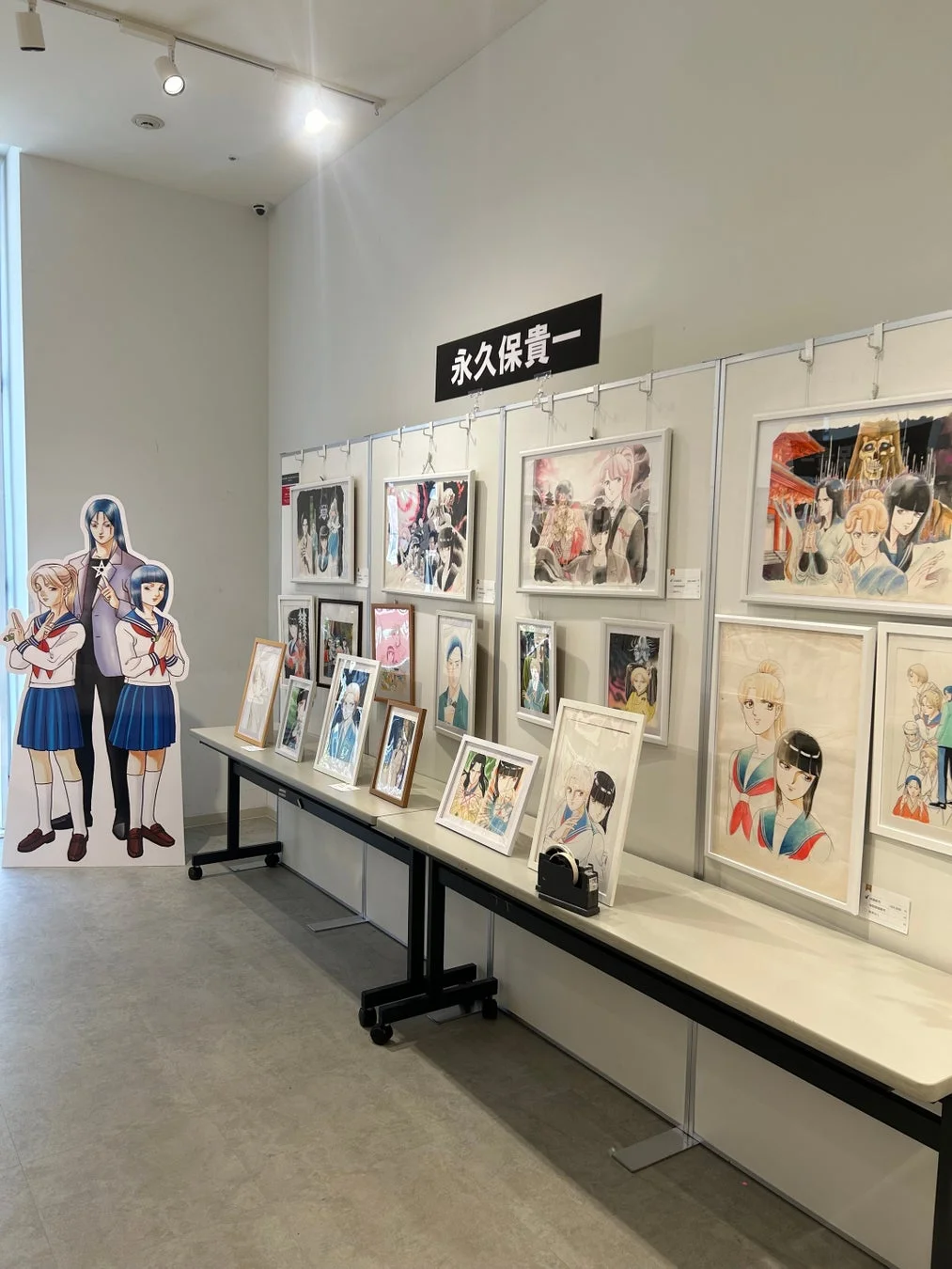 漫画家・永久保貴一のイラスト展示会の様子。壁やテーブルに多数のイラストが飾られ、キャラクターの等身大パネルも設置されている。