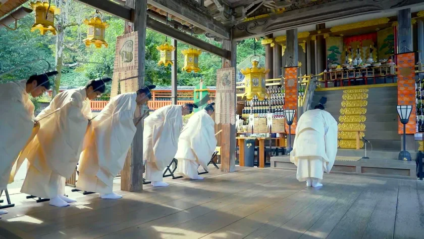 日本の神社で行われている伝統的な神事の様子。白い装束をまとった複数の人々が一列に並び、厳かに頭を下げている。背景には華やかな装飾や供物が並び、格式高い雰囲気が伝わる。
