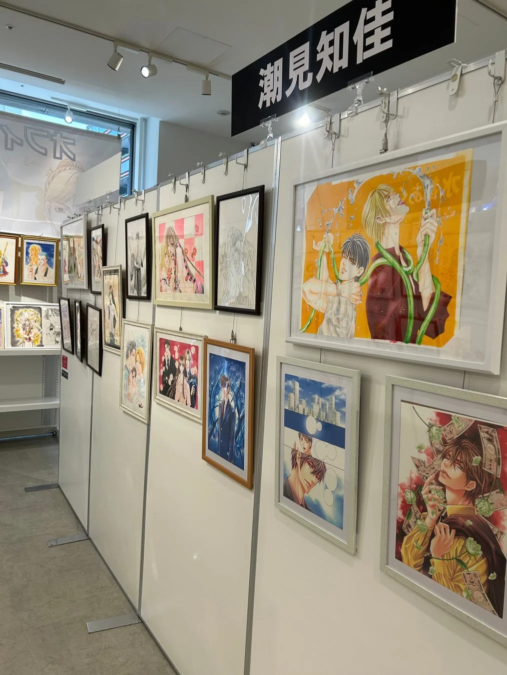 漫画家・潮見知佳のイラスト展の様子を捉えた画像です。白い壁に多数のフレーム入りイラストが飾られ、美しい作品群が並ぶ展示空間が広がっています。