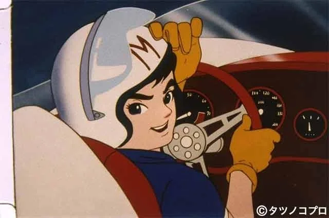 白いヘルメットをかぶり、黄色いグローブをはめて車のハンドルを握るアニメキャラクター「スピードレーサー」の姿。笑顔で前方を見ている。