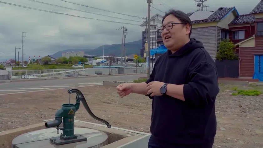 曇り空の下、屋外で緑色の井戸ポンプのそばに立つ男性。背景には日本の街並みと山々が広がり、男性は何かを話している様子です。