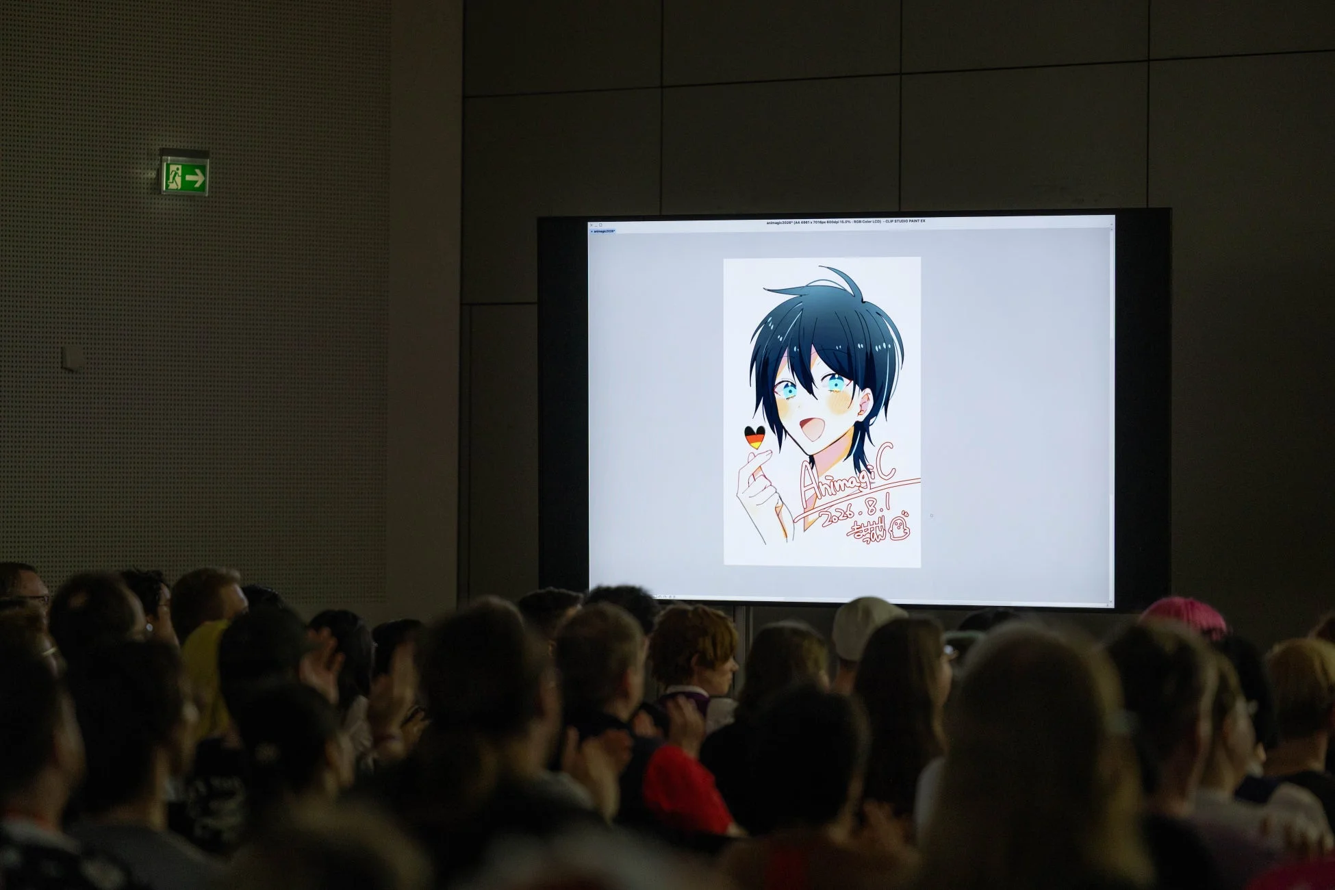 暗い会場で、大きなスクリーンにアニメ風のキャラクターイラストが映し出されています。イラストにはドイツ国旗のハートと「AnimagiC 2023.8.1 お疲れ様」の文字が見え、多くの観客がスクリーンを見ています。