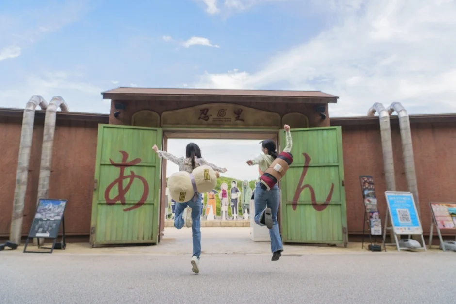 「NARUTO & BORUTO 忍里」のゲート前で、二人の女性がキャラクターの小道具を背負い、楽しそうにジャンプしている様子。テーマパークで友人と過ごす楽しいひとときが伝わります。